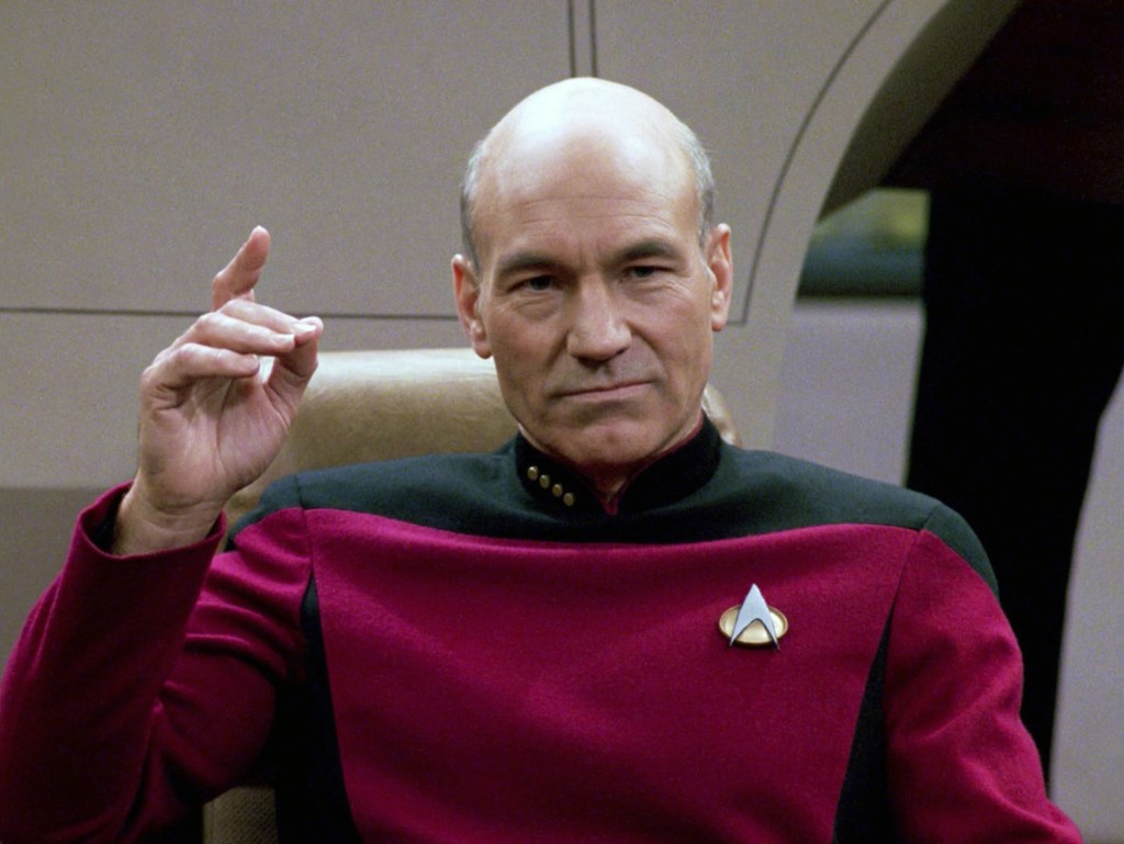 Jean-Luc Picard - a bald man with a stern visage in a maroon captain's uniform, starfleet comms badge on the right, 4 pins on a black collar, his hand raised to order 'make it so'